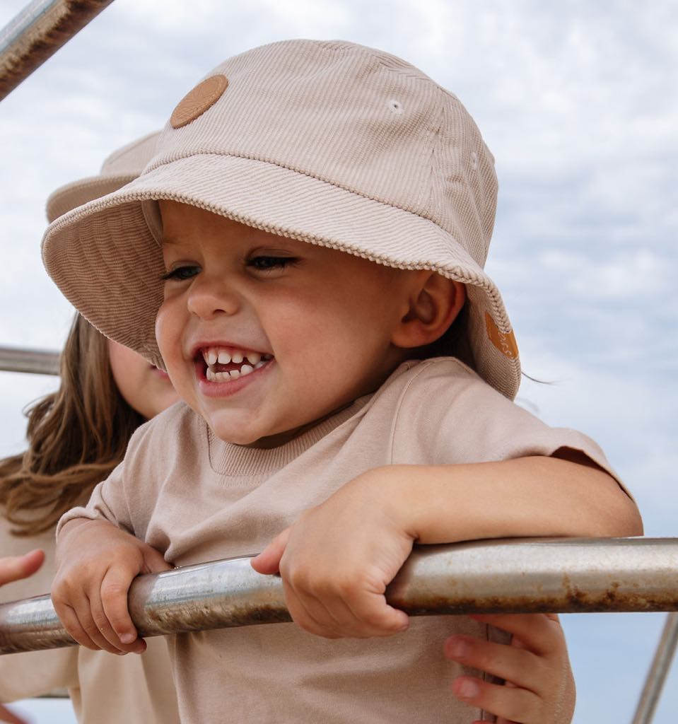 toddler boy wearing a cream corduroy bucket hat handing onto a lifeguard tower at the beach smiling